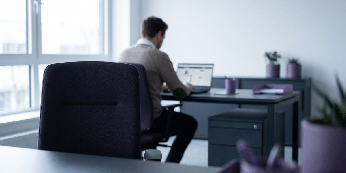 Vista traseira de um profissional trabalhando em um notebook em um escritório moderno, com elementos de decoração em tons de roxo e uma cadeira em primeiro plano.