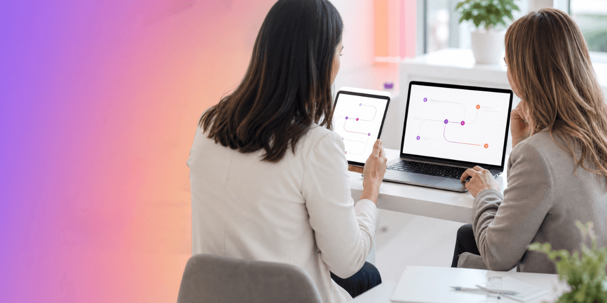 Duas mulheres sentadas em um escritório colaboram olhando para um tablet e um notebook que exibem fluxogramas.